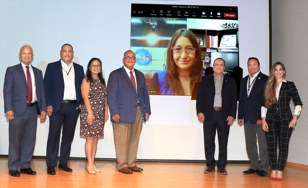 Seven people in business attire stand in front of a monitor with a person on a video call in the background.